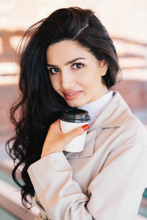 Portrait of fashionable woman with brunette female having charming eyes, pure skin, full lips drinking delicious tea from disposable paper cup resting outdoors enjoying good weather and fresh airの写真素材