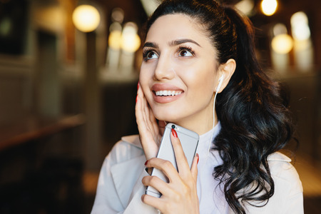 Fashion lifestyle concept. Portrait of cute woman with pony tail having charming smile listening to music with earphones using mobile phone while resting in cafe. Caucasian brunette female with mobileの写真素材