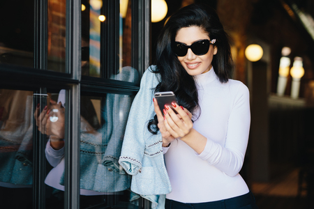 Stylish woman with dark wavy hair dressed in white blouse with jacket wearing big sunglasses holding smartphone communicating with her boyfriend online. Brunette woman updating new applicationsの写真素材