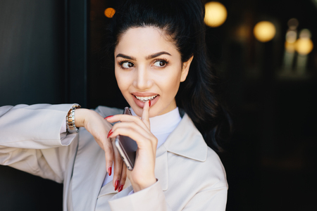Adorable dark-haired woman with appealing eyes, make-up, well-shaped lips and pure skin having red manicure holding her finger on white teeth holding smartphone in hand looking mysteriously asideの写真素材