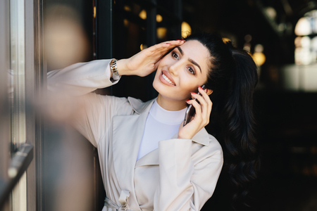 Pretty brunette woman with delicate hands, alluring appearance communicating over cell phone while standing indoors. Young brunette businesswoman having rest speaking with her boyfriend using mobileの写真素材