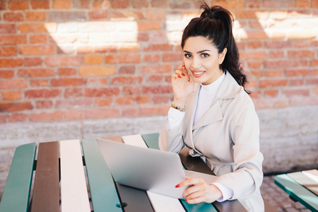 Succsessful businesswoman with dark hair, nice eyes, healthy skin, well-shaped lips having red long nails wearing formal clothes while sitting at cafe using laptop for online communication and workの写真素材