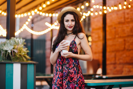 Lovely brunette female in long summer dress and hat, holding in hand paper cup of coffee looking confidently into camera while posing at cozy restaurant or outdoor terrace. Relaxation conceptの写真素材