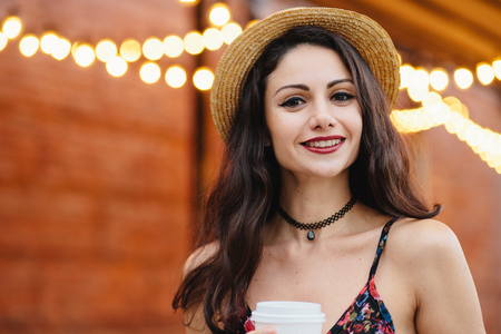 Young female model with dark long hair, hazel eyes and red lips, wearing necklace, summer hat and dress having delightful expression while posing at outdoor cafe. People, rest, emotions conceptの写真素材