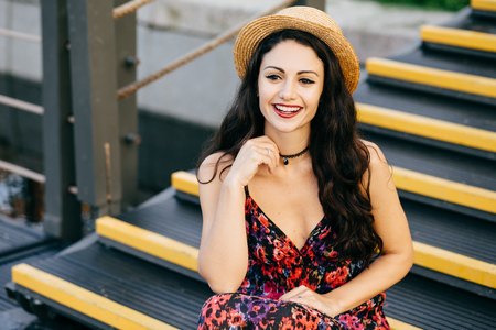 Smiling female with dark luxurious hair wearing dress and straw hat sitting at stairs looking with delightful look into distance. Restful young woman with appealing appearance sitting outdoorsの写真素材