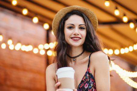 Brunette young lady in summer straw hat and dress, having make-up, holding paper cup with hot drink, having rest at outdoor cafeteria, posing at camera with smile, demonstrating her white teeth.の写真素材