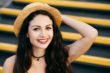 Cheerful woman with dark hair, healthy skin, bright eyes wearing straw hat on head sitting at stairs keeping her hand on head smiling broadly at camera. Carefree female resting alone. Emotions conceptの写真素材