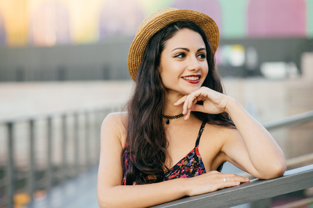 Young female model with long dark thick hair standing at bridge having dreamy expression looking into distance, wearing summer hat and dress resting outdoors. People, relaxation, beauty conceptの写真素材