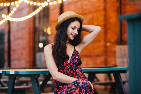 Young attractive woman with European appearance sitting at bench in outdoor cafe, wearing hat and dress, keeping her hand behind head, looking down with delightful expression feeling relaxationの写真素材