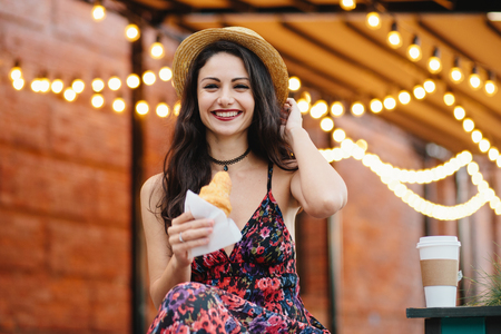 Beautiful female with dark hair, appealing appearance, wearing hat, dress and necklace, holding delicious croissant having cheerful look. Happy young pretty woman having breakfast at terraceの写真素材