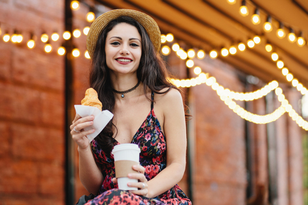 Young female model with dark eyes, red painted lips wearing summer dress and hat holding in hands takeaway coffe and croissant, having breakfast at outdoor restaurant. Food and relaxation conceptの写真素材
