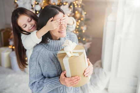 Small adorable female kid prepares surprise for her mother, closes eyes and gives wrapped present as stand at New Year background. Pretty female recieves gift from daughter. Surprisment conceptの写真素材