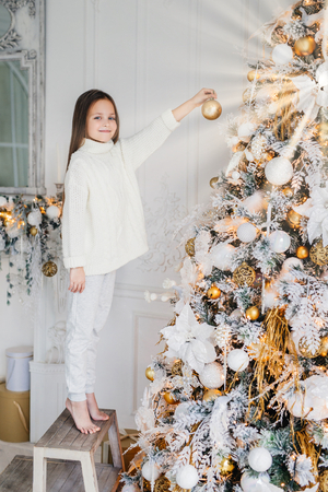 Vertical portrait of female child stands near New Year tree, holds decorated new year ball, decorates fir tree, has happy expression, anticipates miracle. Family, Christmas, holiday, childrenの写真素材