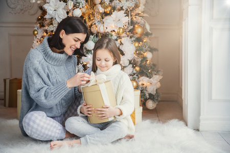 Portrait of pretty girl and beautiful female mother sit together on warm carpet, hold present box, enjoy decorated New Year tree. Family spend time together. Celebration and holidays conceptの写真素材