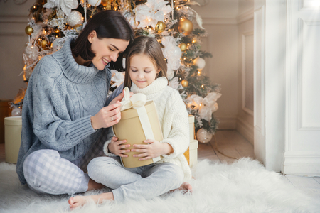 Indoor shot of adorable girl and her mother sit crossed legs, open wrapped present box, have intrigue what is there, sit against adorned New Year or Christmas tree, have sincere gentle smiles on facesの写真素材