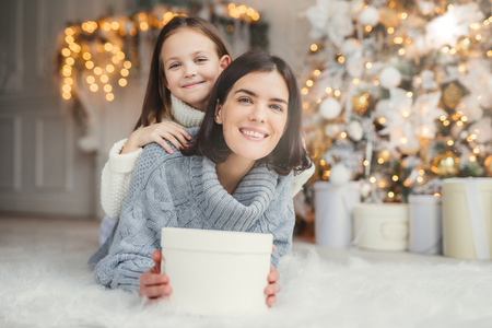 Indoor shot of mother and daughter have fun together, share presents, being in room decorated with garlands and Christmas tree, have joyful expressions, enjoy weekends and winter holidaysの写真素材
