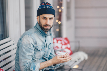 Middle aged bearded male with blue eyes, thick beard and mustache, wears stylish hat and denim jacket, holds cup of hot tea or coffee, has thoughtful expression, thinks about something importantの写真素材