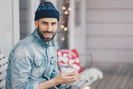 Positive bearded stylish male with pleasant smile, wears fashionable hat and jean jacket, holds cup of aromatic coffee, spends free time at balcony, breathes fresh air, thinks about somethingの写真素材