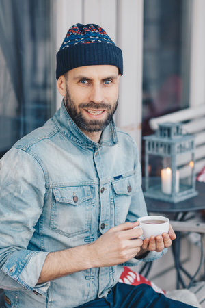 Vertical shot of attractive blue eyed bearded male in stylish clothing, drinks hot tea, looks with pleased expression at camera, spends recreation time at terrace cafe, smiles gently at cameraの写真素材