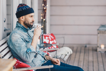 Sideways shot of fashionable bearded male drinks hot tea, enjoys calm atmosphere at home, being deep in thoughts, sits at terrace, enjoys leisure. Handsome middle aged man with beverage. Lifestyleの写真素材