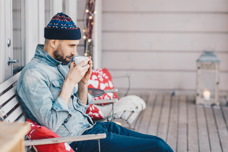 Thoughtful handsome male with beard and mustache, drinks hot tea, warms after stroll, wears fashionable clothing. Pensive man student plans how to pass all exams successfully. Calm atmosphereの写真素材