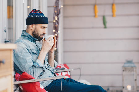 Photo of handsome male remembers something in life while drinks tea or coffee, has thoughtfull look down, recreats at terrace, wears fashionable hat and jean jacket. Leisure and people conceptの写真素材