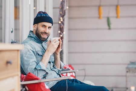 Positive glad attractive male model holds cup of tea, has good rest at balcony, breathes fresh air and enjoys sunny weather, has pleasant smile on face, feels delighted and pleased. People and drinksの写真素材