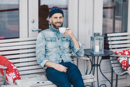 Happy blue eyed male with positive look, sits against cozy interior at terrace cafeteria, enjoys aromatic coffee, has spare time after work. Handsome man has beard, looks joyfully at camera.の写真素材