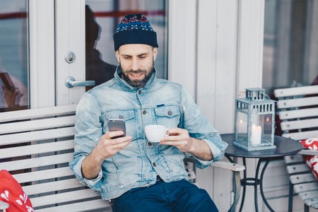 People, leisure and technology concept. Positive bearded young male recreats at terrace cafe, holds modern smart phone, messages online with friends, drinks tea or coffee, wears denim jacketの写真素材