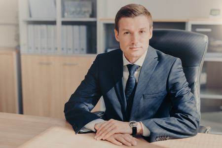 Professional male banker sits in cabinet, wears formal clothes, has confident expression while gives interview concerning banking, poses at office. Man employee arranges meeting of colleaguesの写真素材