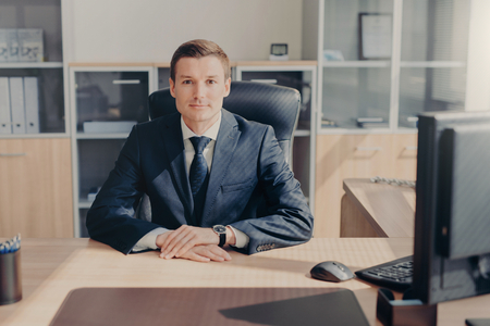 Proud handsome male entrepreneur sits in his cozy cabinet, works on computer, satisfied with successful working day, dressed formally, looks directly into camera. Intelligent employee in officeの写真素材