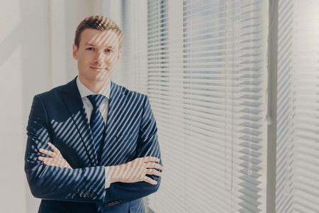Attractive businessman stands arms crossed near office window with jalousie, dressed in luxury elegant clothes, rests after meeting with partner, has satisfied expression. Business lifestyleの写真素材
