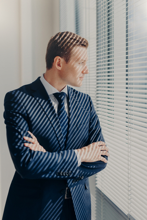 Waist up portrait of handsome male financier looks attentively in office window, has thoughtful expression, rests after conference, has work break, keeps hands crossed, wears elegant clothesの写真素材