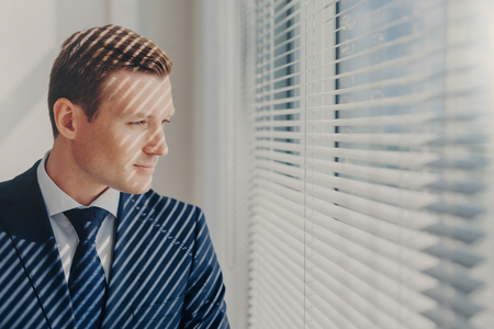Indoor shot of pensive male economist dressed in formal outfit, looks pensively at window with jalousie conemplates about work during dinner break feels tired after working. Business, people, thoughtsの写真素材