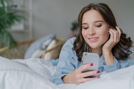 Beautiful woman listens music, makes video call, lies in comfortable bed in morning, dressed casually, has charming smile, poses in bedroom. People, relaxation and modern technologies conceptの写真素材