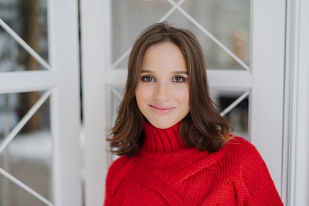Headshot of charming woman with dark hair, wears knitted sweater, looks directly at camera, has...