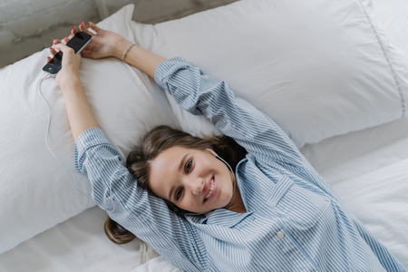 Relaxed European woman with toothy enjoys good rest in bed, stretches during morning, holds modern cell phone, listens favourite song with earphones, rejoices start new day. Horizontal view.の写真素材