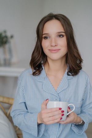 Indoor shot of dreamy young European woman with charming look, dressed in striped pyjamas, drinks coffee, thinks about something, enjoys calm domestic atmosphere. People, drinking and home conceptの写真素材