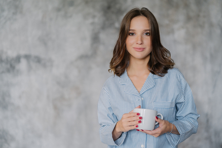 Attractive woman dressed in nightwear, holds white mug with coffee or tea, has morning drink, poses indoor against blurred background with copy space for your advertisement or promotional contentの写真素材