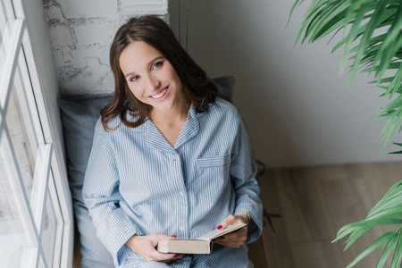Photo of cheerful woman reads romantic story, holds book, wears casual clothes, poses on window sill in cozy room. View from above. Satisfied female model being in good mood, reads textbook.の写真素材