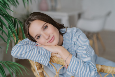 Headshot of brunette young woman with healthy skin and make up, leans on hands, feels lonely while being at home, poses against blurred backround. Relaxed girlfriend in striped blue costume.の写真素材