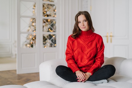 Horizontal shot of relaxed housewife dressed in warm red knitted sweater, sits crossed legs in lotus pose on couch, in spaciouss room with New Year tree in background, light colours. Rest conceptの写真素材
