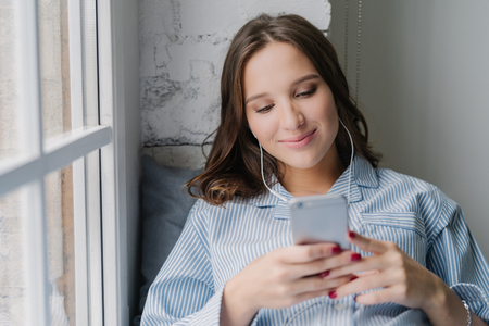 Photo of Cauasian woman selects favourite song in playlist, uses contemporary cellular and high quality earphones, dressed in casual outfit, poses on window sill in cozy room alone. Technologyの写真素材