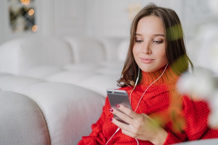 Beautiful young woman holds cell phone, listens music from playlist, surfes social networks and messages with friends, enjoys calm atmosphere at home, sits on comfortable sofa. Lifestyle conceptの写真素材