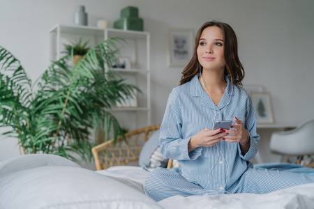 Shot of pensive female model uses smart phone and earphones for watching movie, connected to wireless internet, dressed in striped casual nightwear, poses in lotus pose on bed, has good restの写真素材