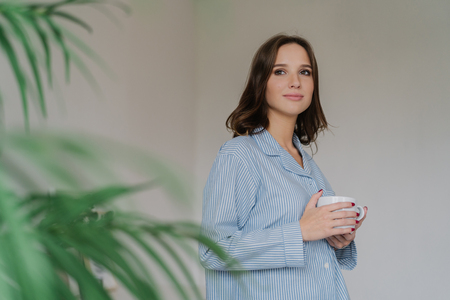 Photo of thoughtful young woman with dark hair, appealing appearance, warms with hot beverage, dressed in domestic costume, poses indoor near green plant, blank space for your advertisement.の写真素材