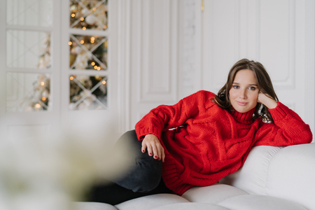 Indoor shot of pretty dark haired woman in warm winter clothes, leans at hand, lies on comfortable white sofa, looks directly at camera with calm expression. Domestic atmosphere. Time at homeの写真素材