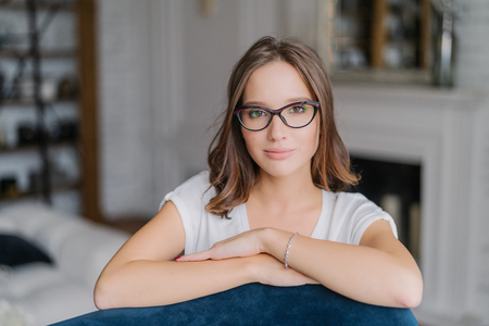 Indoor view of pretty woman in spectacles, keeps hands on back of sofa, poses in living room at home, thinks about something, dressed in casual clothes. People, free time and lifestyle conceptの写真素材