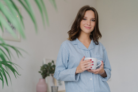 Waist up shot of pleasant looking young woman dressed in striped pyjamas, drinks hot beverage after awakening, has sparre time at home, stands indoor against cozy interior. People and rest conceptの写真素材
