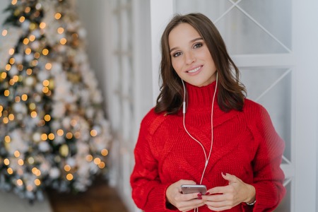 Cheerful good looking woman has toothy smile, enjoys nice track in earphones, updates playlist on smart phone, dressed in warm knitted sweater, poses against decorated Christmas tree in backgroundの写真素材